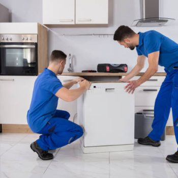 Two Young Male Movers In Uniform Placing Dishwasher In Kitchen